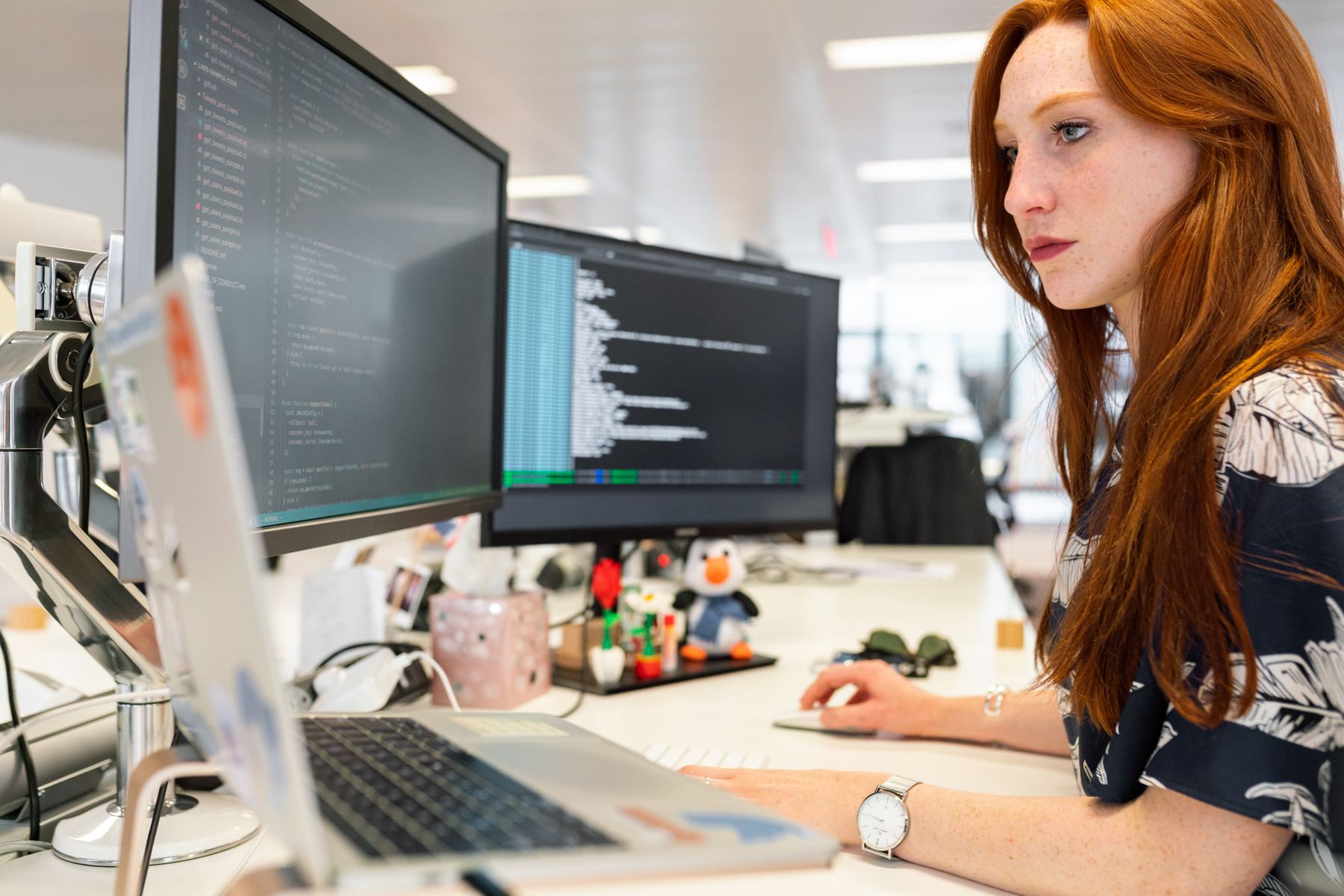 Software engineer reviewing code across two monitors in an open plan technology office